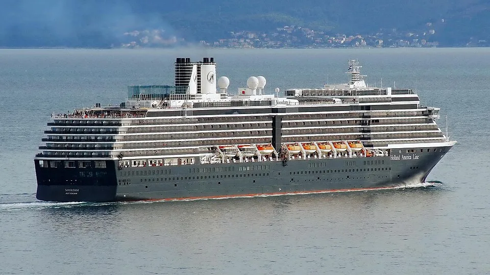 The Holland America Line cruise ship Noordam sailing across open water. The large multi-deck vessel features a dark blue hull, white upper decks, orange lifeboats, and a white funnel with the company logo.