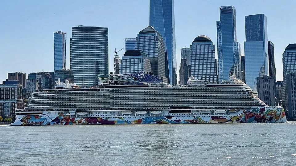 The massive Norwegian Aqua cruise ship sails along a river, showcasing a vibrant, multicolored art design across its lower hull. In the background, the iconic skyscrapers of the Lower Manhattan skyline tower under a clear blue sky.
