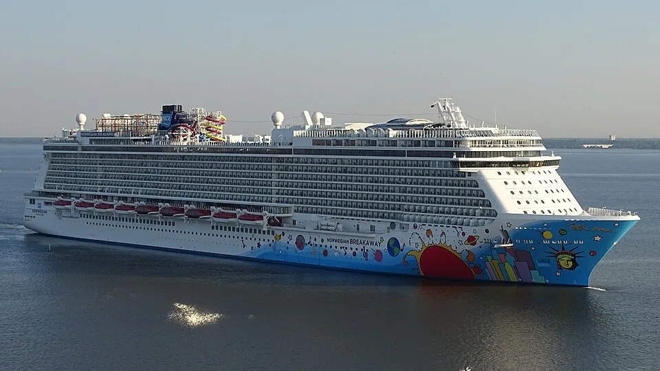 The massive Norwegian Breakaway cruise ship sails on calm water. The white ship has rows of balconies and a colorful hull painted with a bright sun, ocean waves, and New York landmarks. A water park with waterslides is visible on the top deck.
