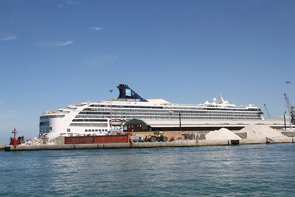 The large white cruise ship Norwegian Spirit is docked at an industrial port terminal. The ship features a dark blue funnel with the NCL logo, while shipping containers, large sand mounds, and construction vehicles sit on the pier in the foreground under a clear blue sky.