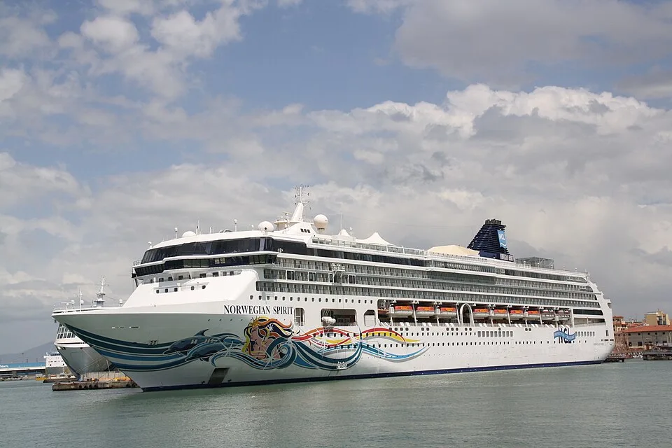 The large cruise ship Norwegian Spirit floats on calm water under a partly cloudy sky. The white vessel features vibrant hull art with waves and a mermaid figure, rows of cabin windows, orange lifeboats, and a dark blue funnel at the back.