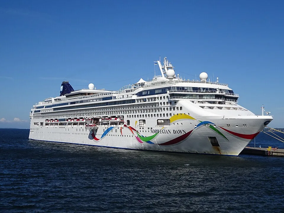 The large cruise ship Norwegian Dawn floats on dark blue water under a clear sky. The white vessel features bright, colorful swirling ribbons painted on its hull, rows of windows, and lifeboats along the side. It is docked at a pier on the right.