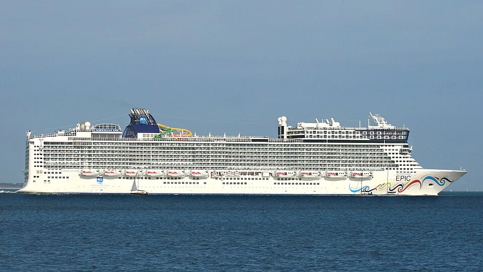The massive Norwegian Epic cruise ship sails across a calm blue sea. The white ship has rows of cabin balconies, a unique boxy forward structure, and a colorful wave design painted on its bow. A small sailboat sits in the water nearby.