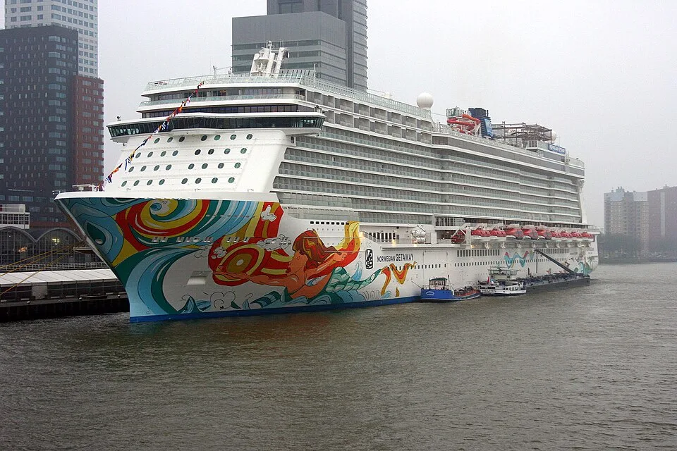 The Norwegian Getaway cruise ship is docked on a river under a foggy sky. The white ship has a colorful mermaid painting on its bow and rows of cabin balconies. Smaller support boats are anchored alongside it, with city buildings visible in the background.