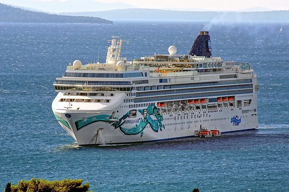 The cruise ship Norwegian Jade sails on wavy blue water against a background of distant hills. The large white vessel features colorful blue and green hull art, multiple window-lined decks, and a tall dark funnel emitting faint white smoke.