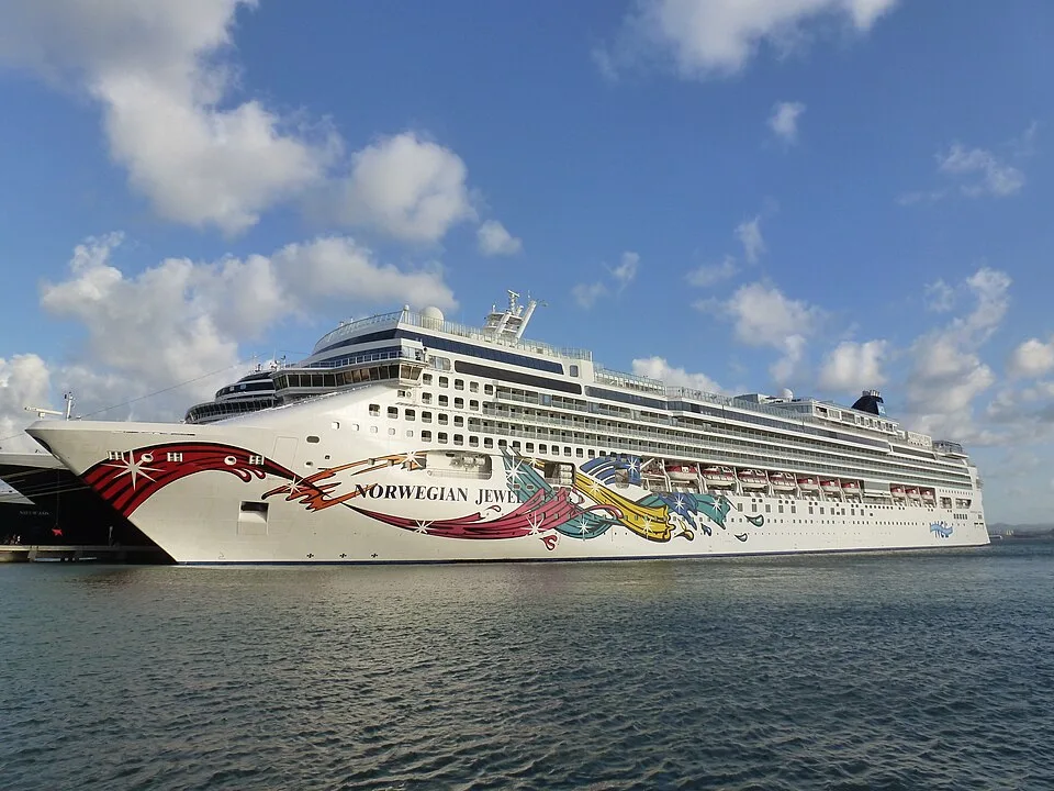 The large cruise ship Norwegian Jewel rests on calm blue water under a partly cloudy sky. The white vessel displays vibrant, multi-colored gemstone artwork streaming along its hull, with multiple passenger decks, rectangular windows, and a dark blue funnel visible near the back.