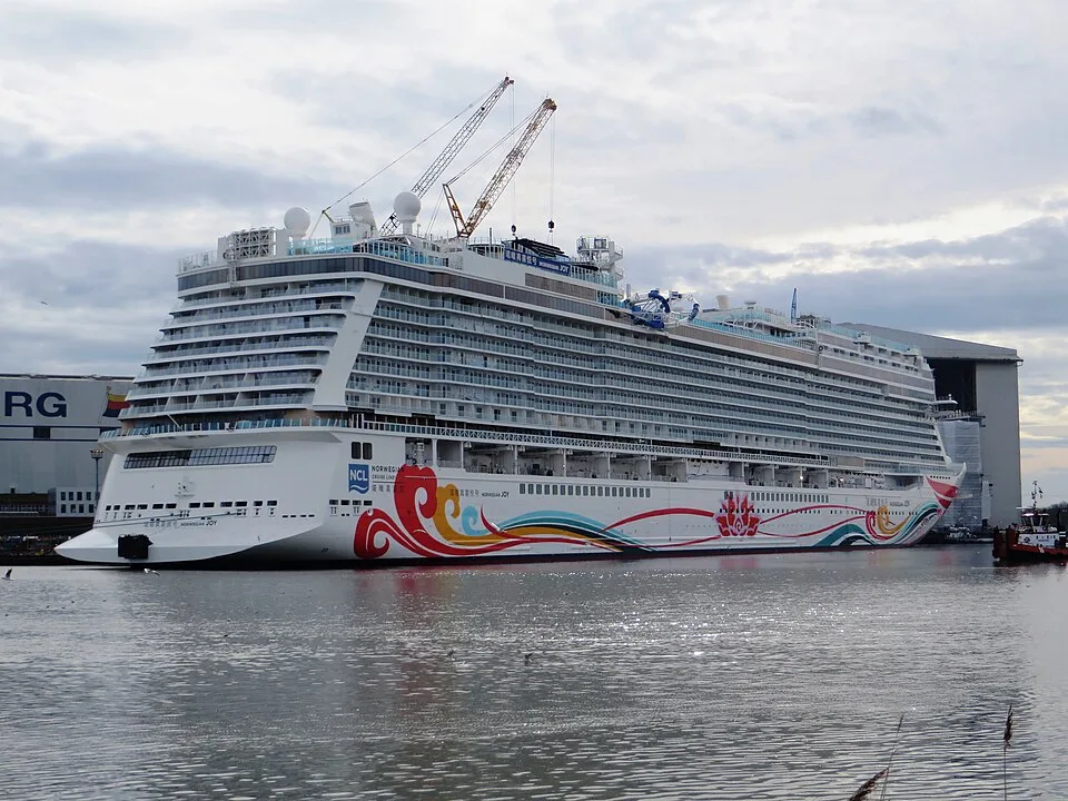 The Norwegian Joy cruise ship is docked in a harbor under a cloudy sky. The massive white vessel features vibrant red, blue, and yellow artwork along its side. Two tall construction cranes reach over its top decks, and large industrial port buildings stand in the background.