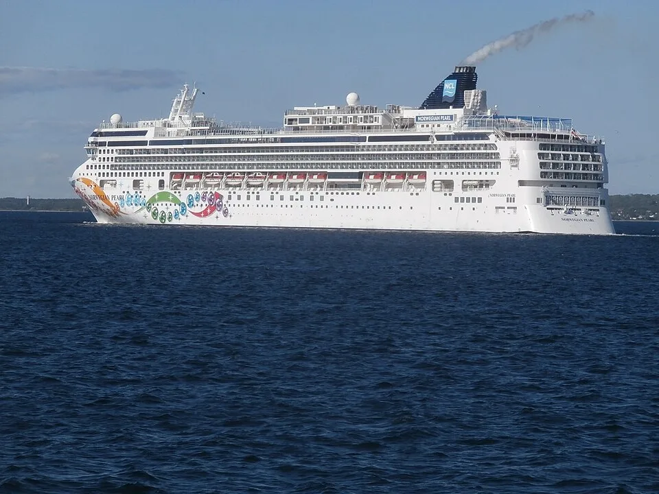 The large cruise ship Norwegian Pearl sails on deep blue water under a clear sky. The white vessel features vibrant, multicolored pearl and ribbon artwork on its hull, orange lifeboats, and a dark blue funnel with the NCL logo emitting a steady trail of gray smoke.