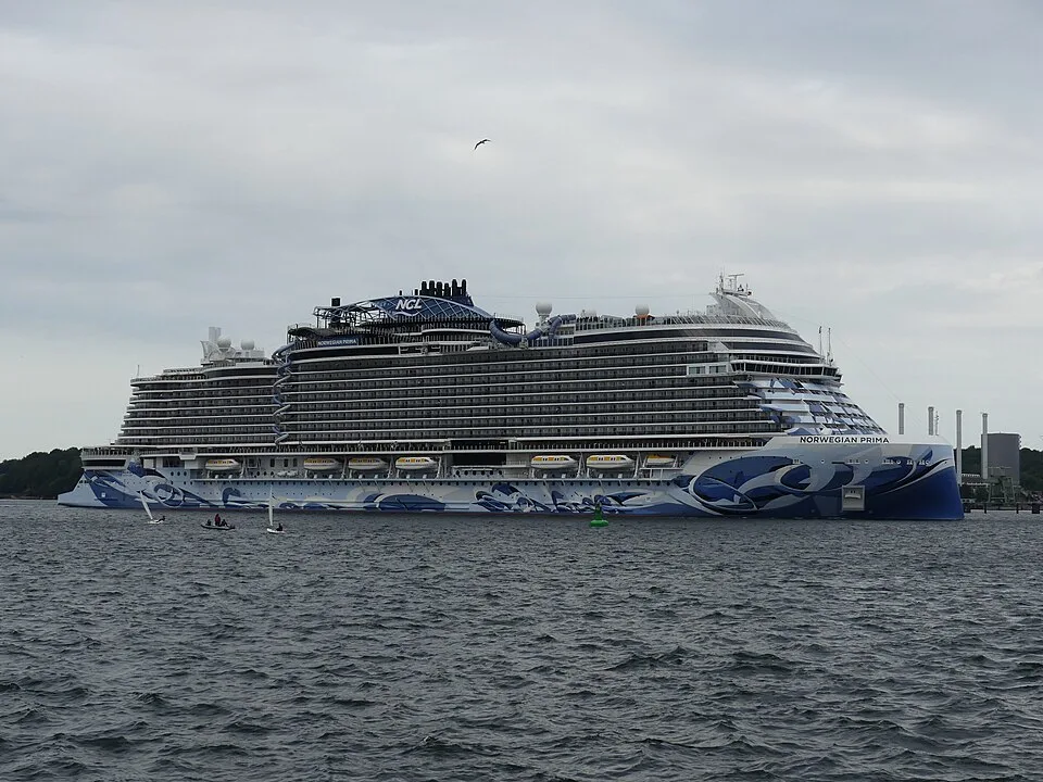 The massive Norwegian Prima cruise ship sails on choppy water under an overcast sky. The white ship has rows of dark balcony cabins and features a striking blue and white abstract hull design. A few small boats float nearby, and a lone bird flies overhead in the distance.