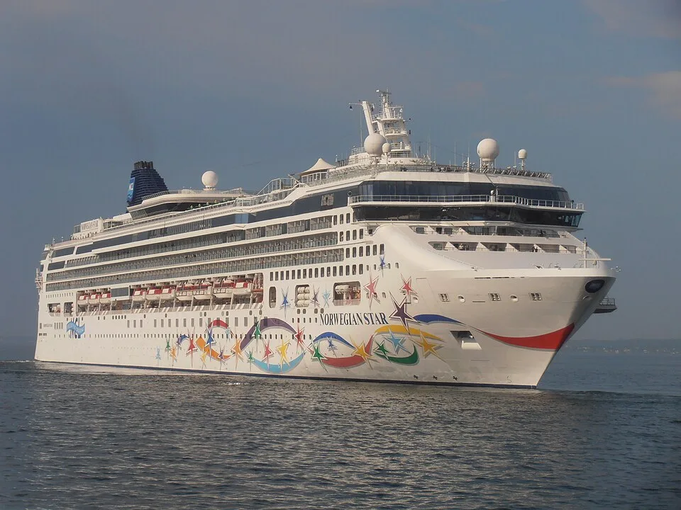 The Norwegian Star cruise ship sails across open water under a clear sky. The white ship has rows of cabin windows and features a colorful pattern of stars and swirling ribbons painted along its side. A dark blue smokestack stands out near the back of the vessel.