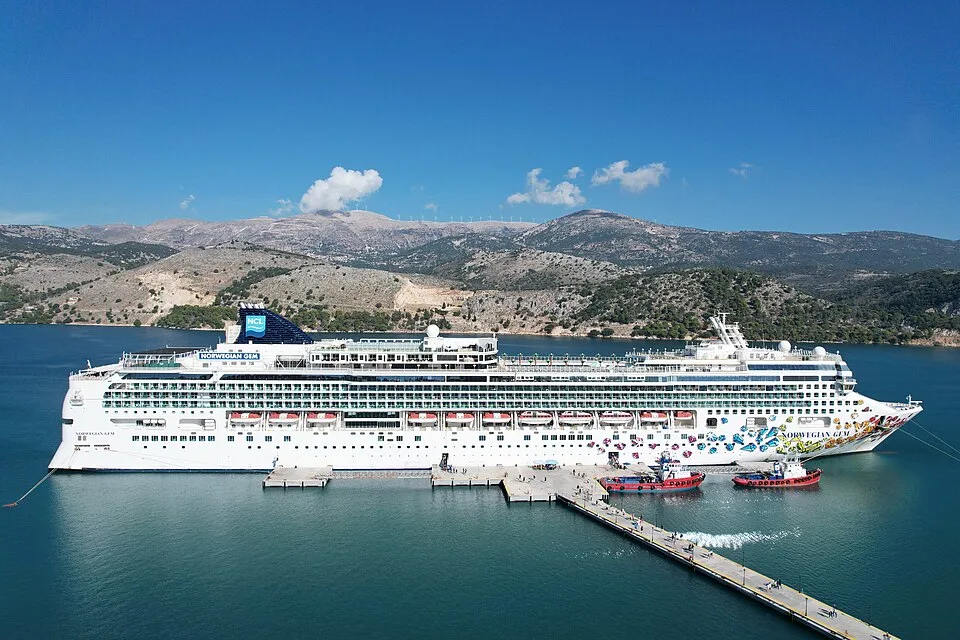 The large white cruise ship Norwegian Gem is docked at a pier in calm turquoise water. The ship features colorful hull artwork, a dark blue funnel with the NCL logo, and orange lifeboats. Two small tugboats rest nearby against a backdrop of arid, rolling mountains under a bright blue sky.