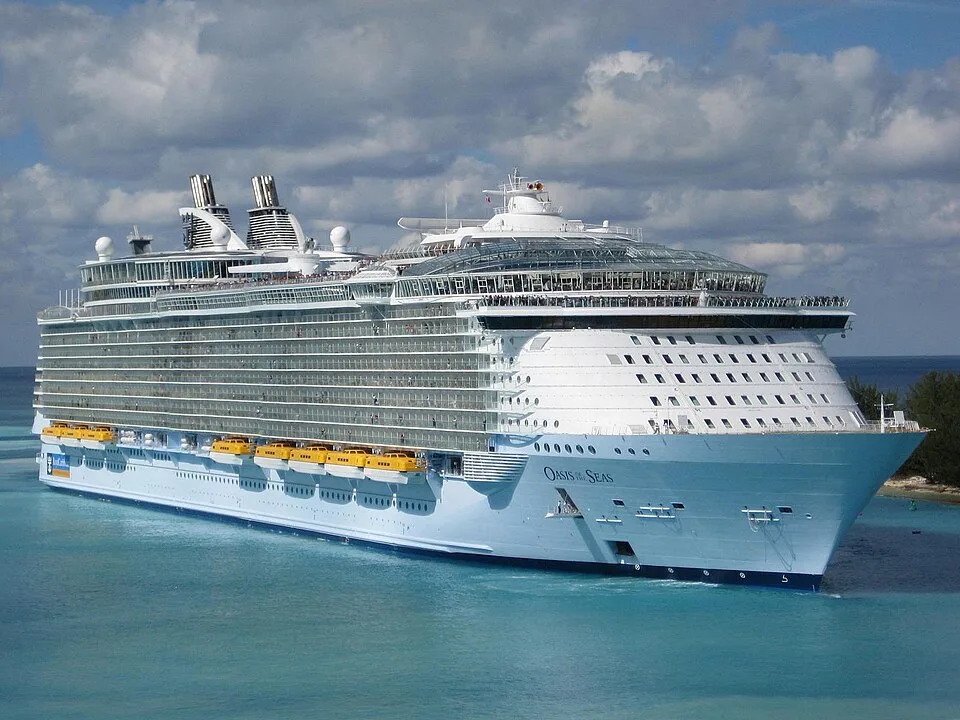 A massive white cruise ship named "Oasis of the Seas" sailing on bright blue water under a cloudy sky. The multi-story ship features rows of balconies, yellow lifeboats along the side, and two large smokestacks near the top.