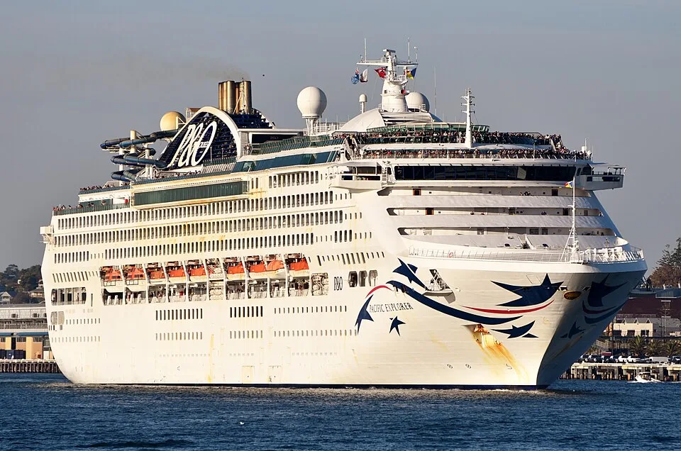 The large P&O cruise ship Pacific Explorer sails on blue water under a clear sky. The white vessel features blue star graphics on its bow, the P&O logo near its gold funnel, multiple decks filled with passengers, and orange lifeboats tucked along its side.
