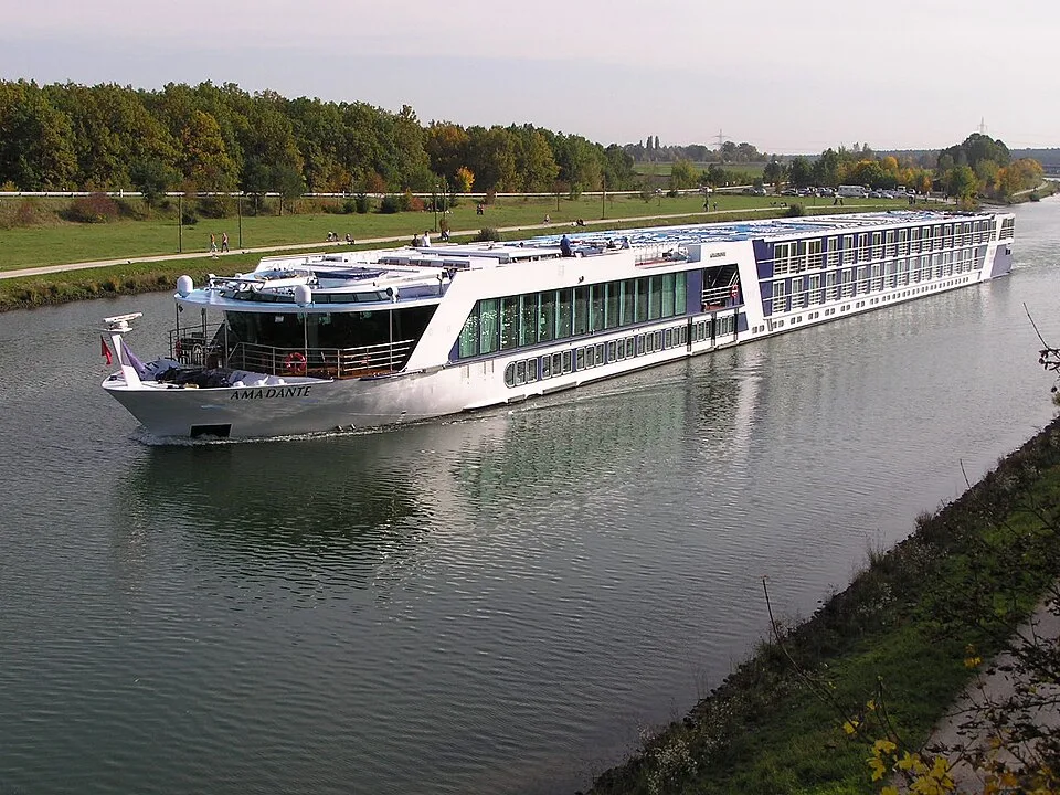 A long, white river cruise ship named "AmaDante" sailing along a calm canal. The modern vessel features a glass-enclosed lounge, multi-tiered cabins with windows, and a flat top deck, with green lawns, trees, and paths visible along the far shoreline.