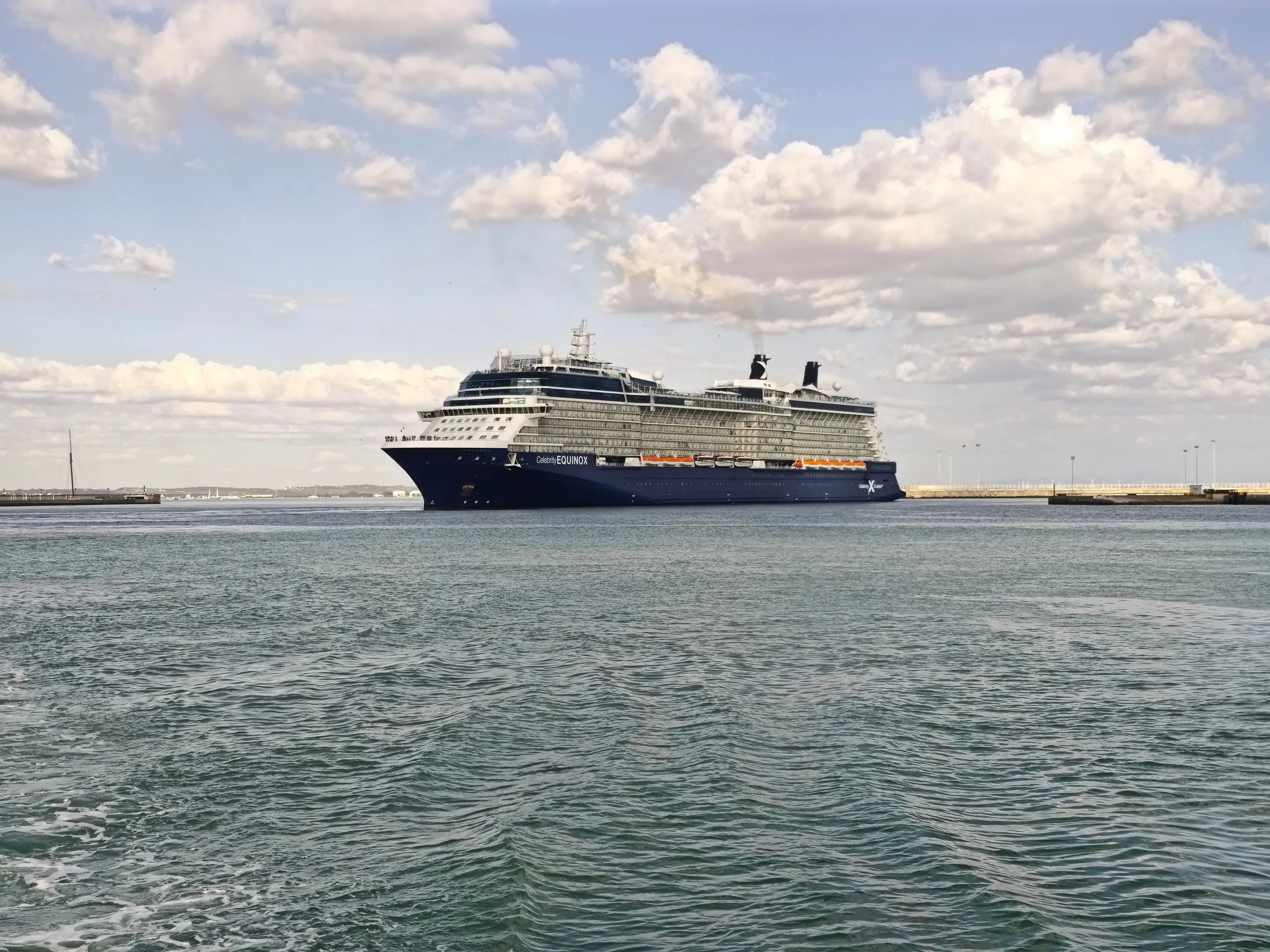 Elegant cruise ship navigates calm waters in a Spanish harbor under a partly cloudy sky.