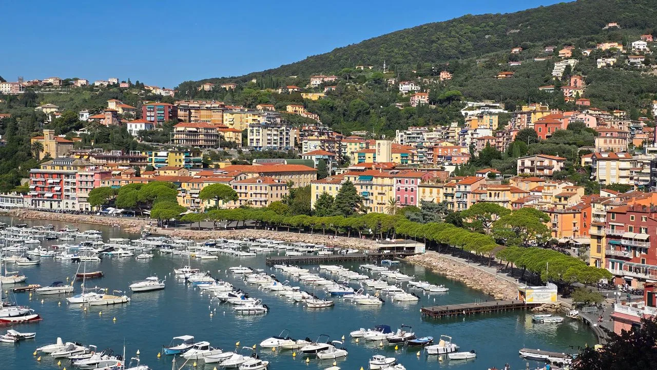 Aerial view of a vibrant Mediterranean coastal town with a marina full of boats, surrounded by lush green hills.