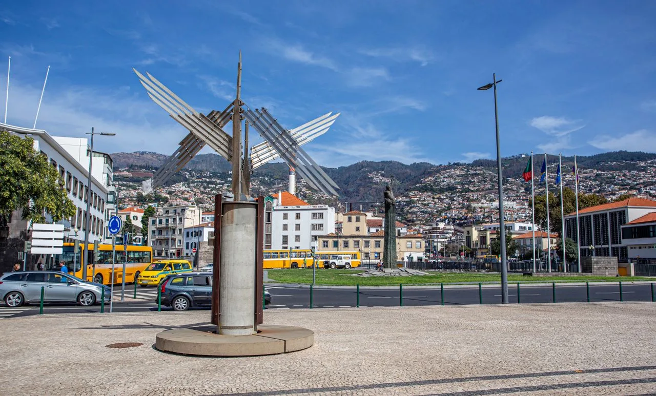 Street view of Funchal cityscape featuring a modern sculpture and mountain backdrop.