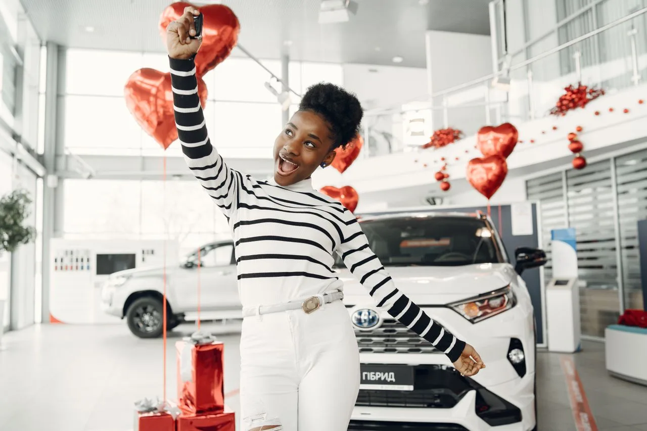 Woman excitedly holds car key in dealership with red balloons, symbolizing celebration and achievement.