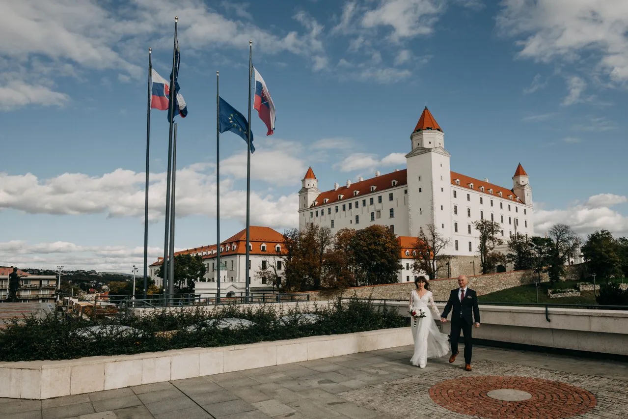 A newlywed couple holding hands in front of Bratislava Castle, Slovakia.