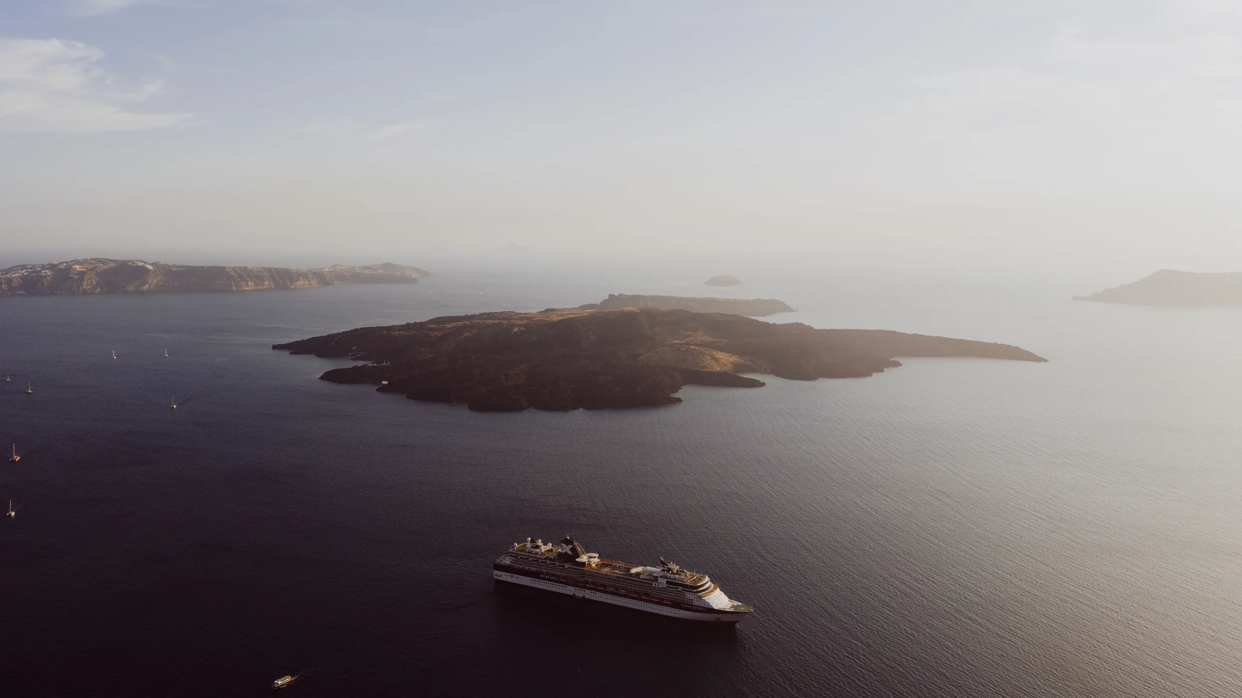 Aerial photo of a cruise ship sailing near Santorini island under a serene sunset sky.