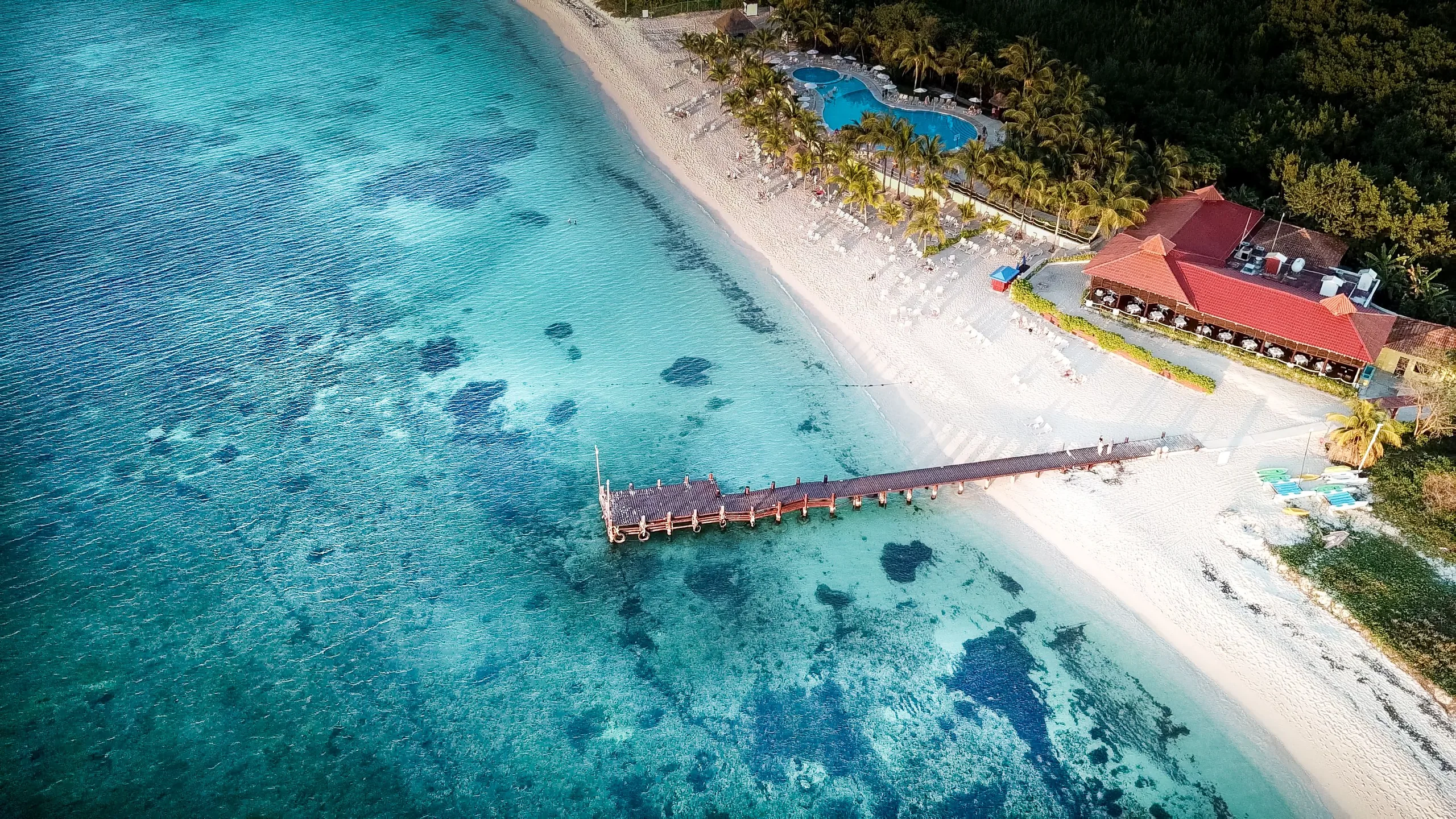 Aerial shot of a tropical beach with a pier, turquoise waters, and a resort in Cozumel.