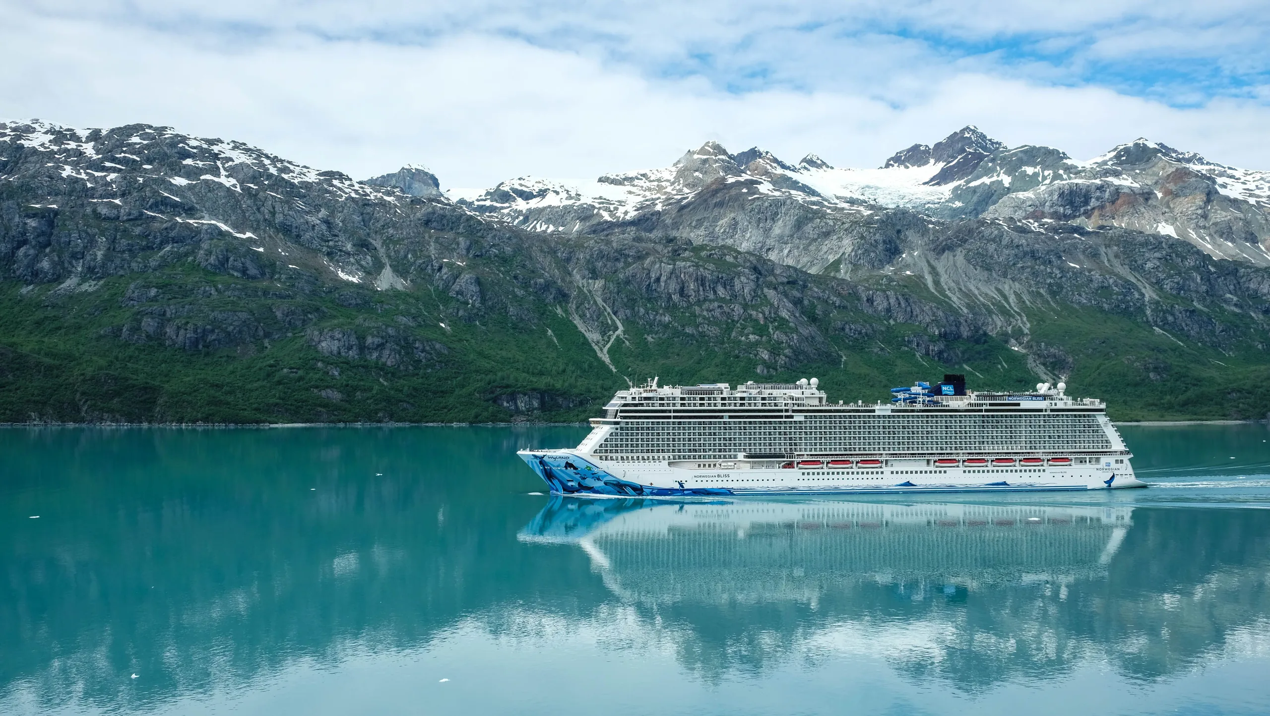 A majestic cruise ship (Norwegian Bliss) sailing through clear waters with snowy mountains as a backdrop.