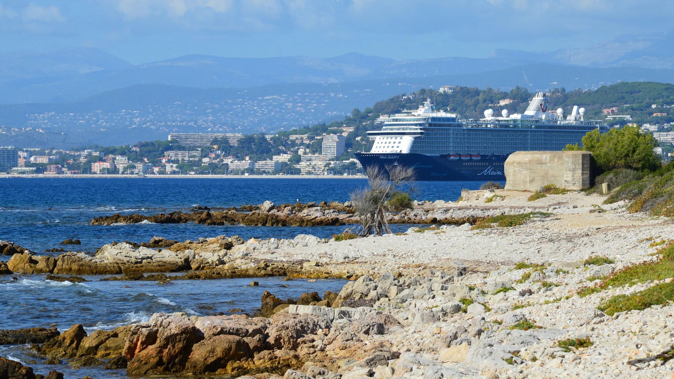 A luxury cruise ship sails along a scenic rocky coastline with a city skyline backdrop.