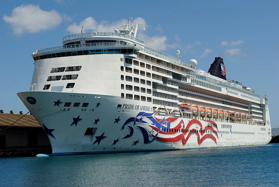 The Pride of America cruise ship sits docked in clear blue water under a sunny sky. The large white vessel features patriotic hull artwork with red and white stripes and blue stars. Its multi-story decks rise above the water, showcasing cabin balconies and a dark funnel.