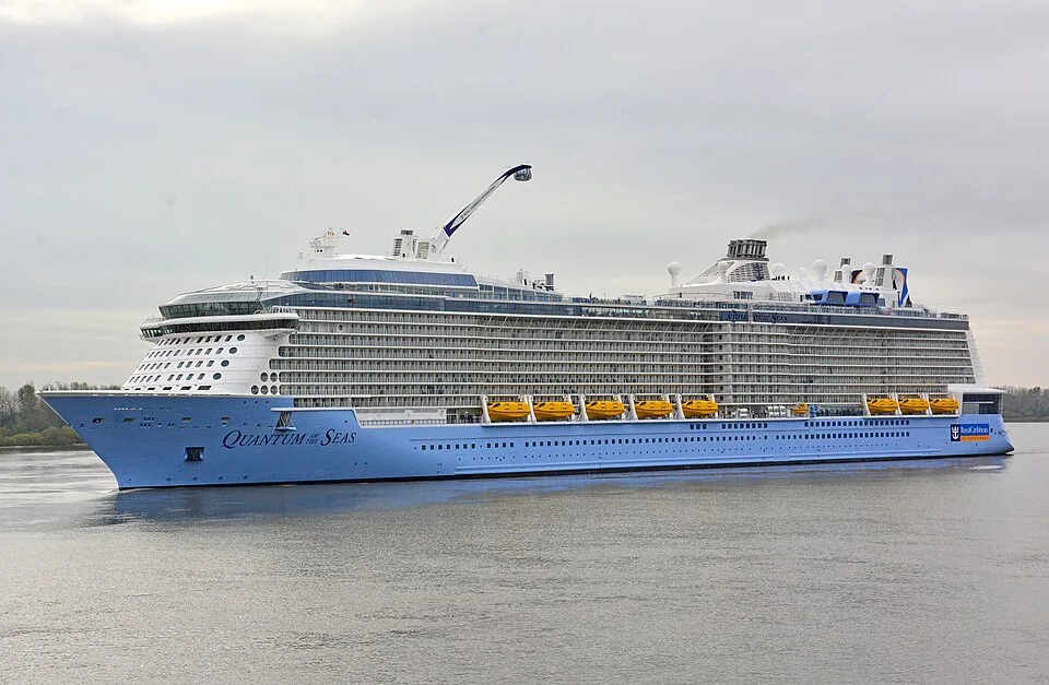 A large Royal Caribbean cruise ship named "Quantum of the Seas" traveling on calm water. The massive light-blue and white ship features many deck levels, yellow lifeboats, a tall mechanical arm with a viewing capsule on top, and a grey sky overhead.