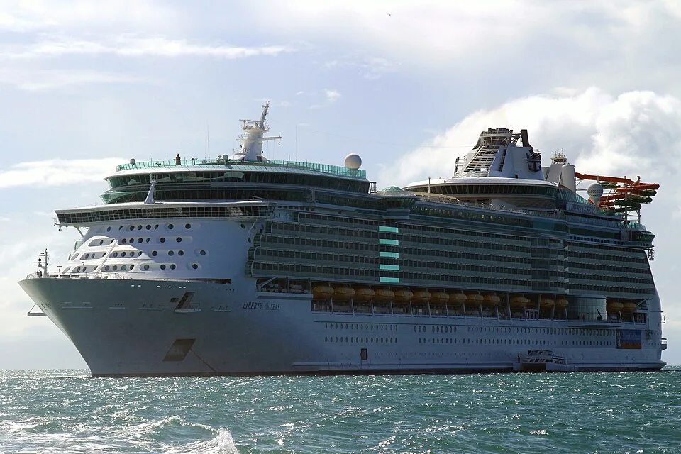 The massive white cruise ship Liberty of the Seas sails on choppy blue-green water under a bright sky with large white clouds. Yellow lifeboats line the side of its multi-deck hull.