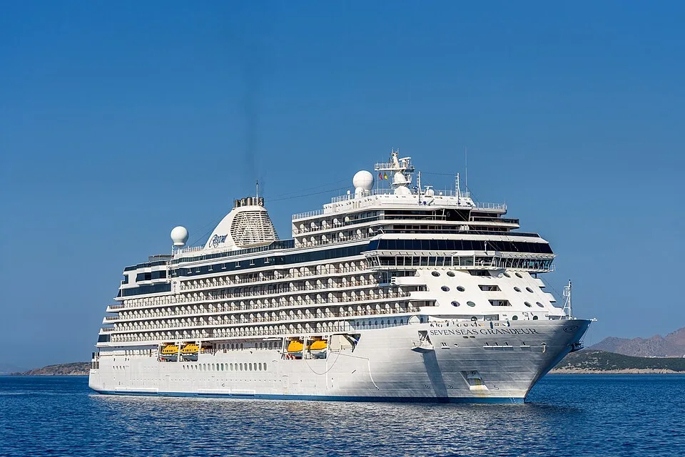 The white luxury cruise ship Seven Seas Grandeur sails on calm blue water under a clear blue sky, with hilly land in the far background.