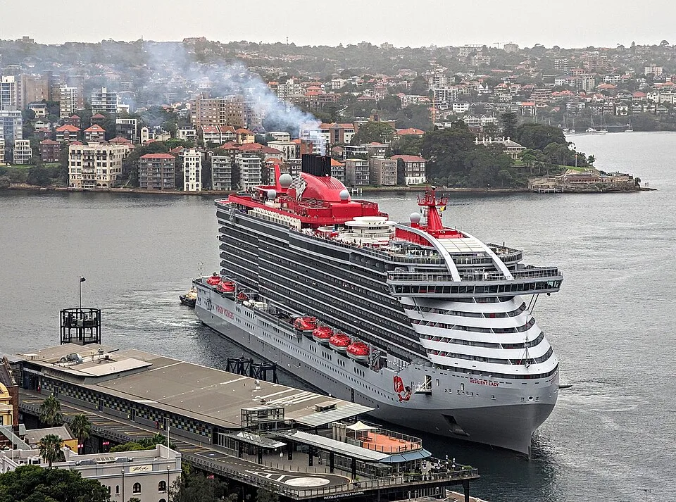 The massive Virgin Voyages cruise ship Resilient Lady is docked at a city terminal. The vessel features a grey hull, towering passenger decks, a red top structure with a smoking black funnel, and red lifeboats. A dense hillside city stands in the background across the water.