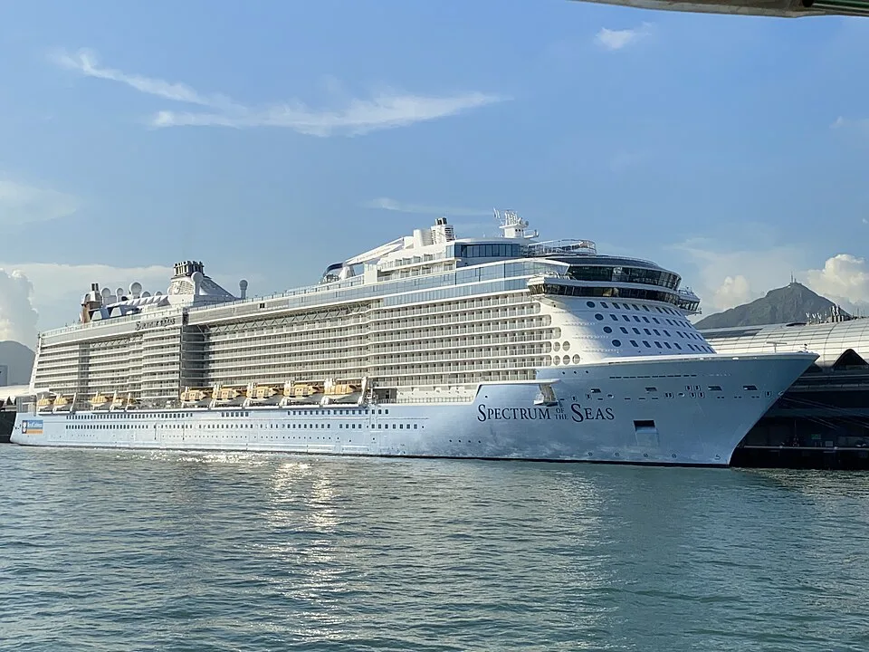 A massive white Royal Caribbean cruise ship, named "Spectrum of the Seas," docked in calm water next to a port terminal. The multi-story ship features numerous balconies, with mountains visible in the hazy background under a partly cloudy sky.