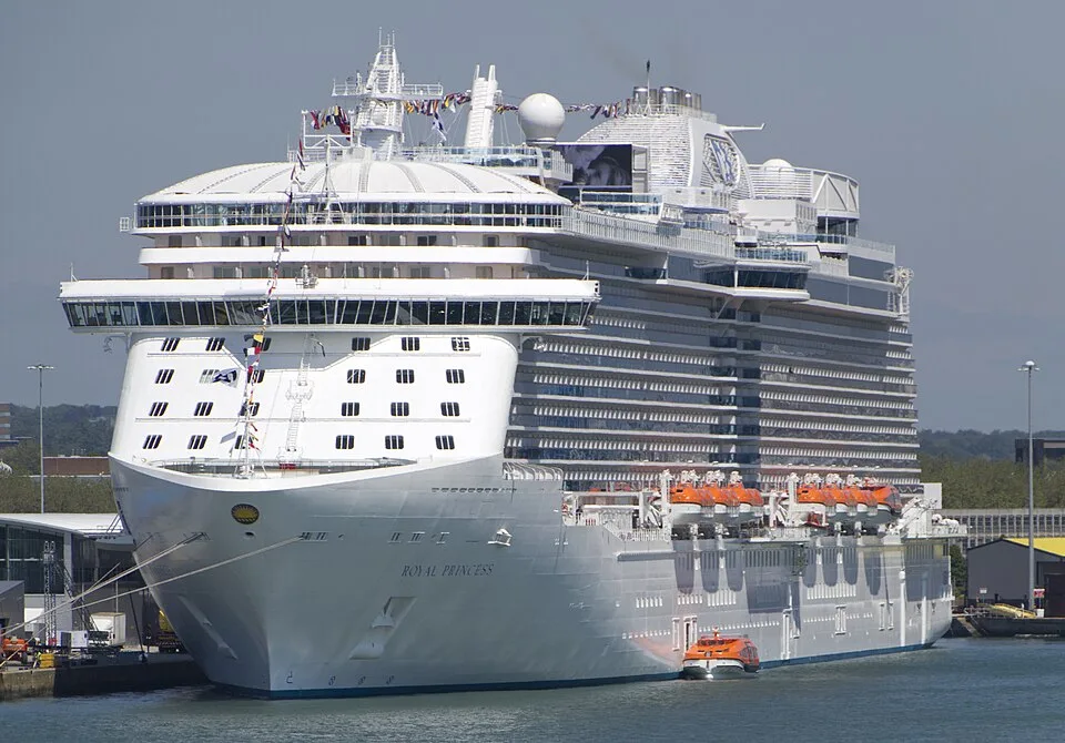 The Royal Princess cruise ship sits docked at a port on a sunny day. The huge white ship features multi-level decks with glass balconies, bright orange lifeboats, and colorful nautical flags strung across the front. A small orange tender boat floats near its side.
