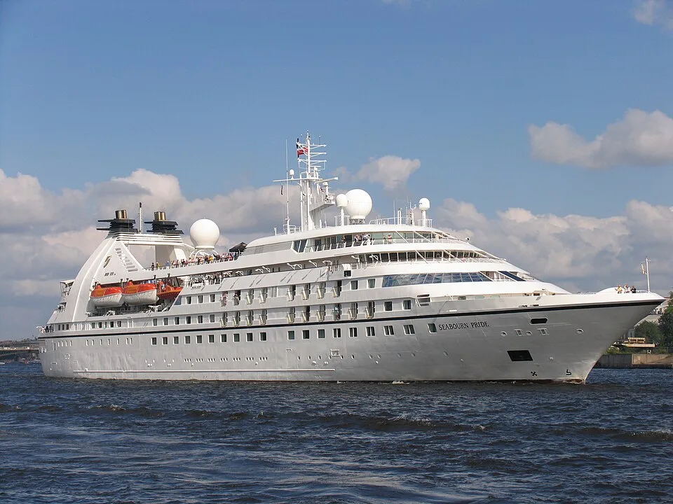 A white luxury cruise ship named "Seabourn Pride" cruising on choppy dark blue water. The multi-deck vessel features white radar domes, orange lifeboats near the stern, and small figures of passengers visible on the upper open-air decks under a cloudy blue sky.