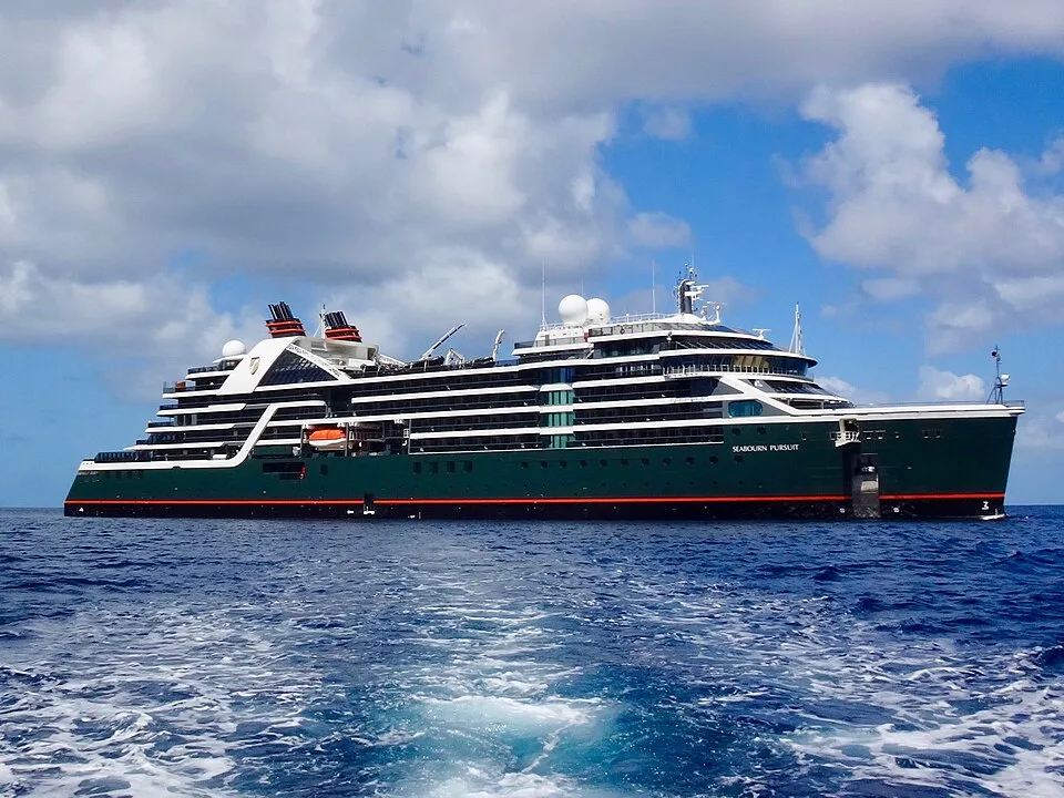 A luxury cruise ship named "Seabourn Pursuit" floating on deep blue water under a partly cloudy sky. The multi-level ship features a distinct dark green hull with an orange stripe, white upper decks, and black funnels with red trim.