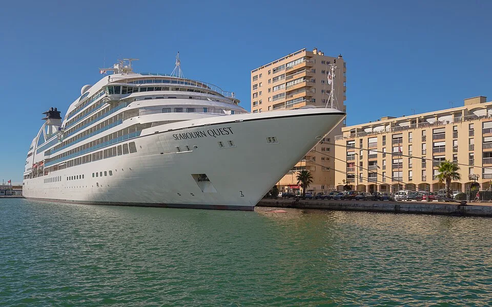 The luxury cruise ship Seabourn Quest is docked at a city port terminal on calm green water. The sleek white vessel features its name near the bow, multiple tiers of windows, and a black and white funnel structure, set against a backdrop of tall residential buildings under a blue sky.