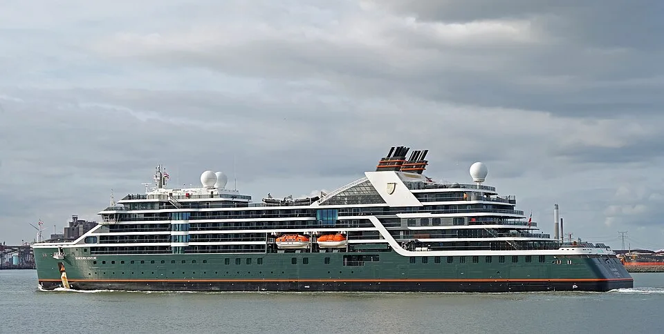 A dark green and white luxury expedition ship named "Seabourn Venture" sailing on calm water. The ship features multi-tiered decks, orange lifeboats tucked along the side, black smokestacks with red accents, and a large white radar dome under an overcast sky.
