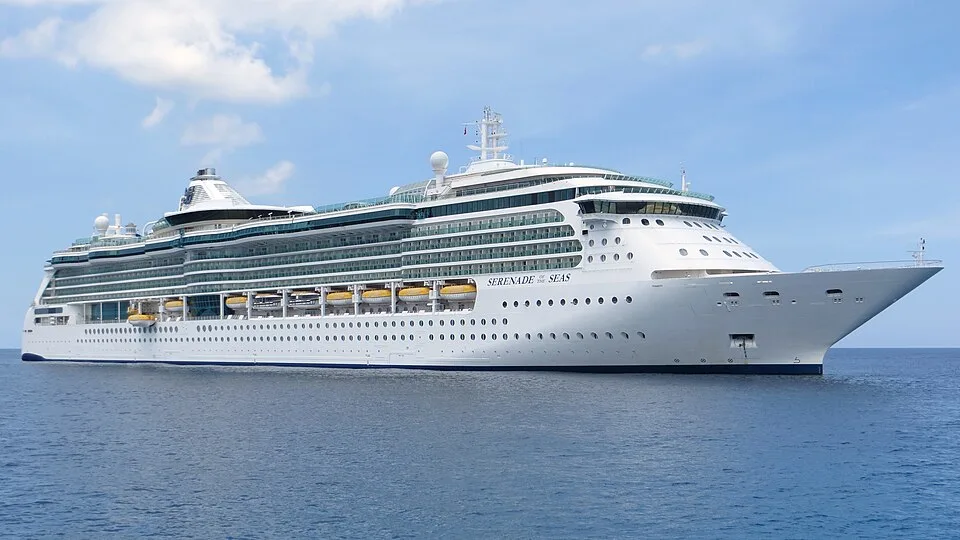 A large white cruise ship named "Serenade of the Seas" floating on calm, dark blue ocean water under a light blue sky with scattered clouds. The ship features multiple decks with glass balconies, yellow lifeboats, and a distinctive dark glass-walled upper deck structure.