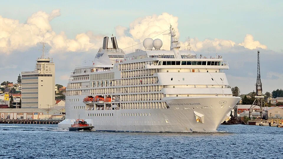 The large white luxury cruise ship Seven Seas Navigator moves through a harbor with a smaller pilot boat alongside it. Buildings and a harbor crane are visible in the background under a cloudy sky.