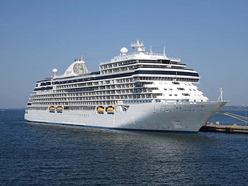 The luxury cruise ship Seven Seas Splendor sits in deep blue water next to a concrete pier. The massive white vessel features its name clearly printed on the bow, multiple tiers of dark-trimmed balconies, yellow lifeboats, and a unique latticed funnel under a clear blue sky.