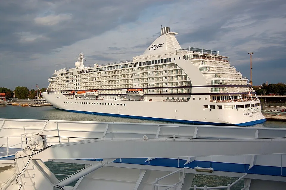 The large white luxury cruise ship Seven Seas Voyager, featuring the Regent logo on its funnel, is docked or sailing near a port under a cloudy sky, viewed from another vessel's deck structure in the foreground.