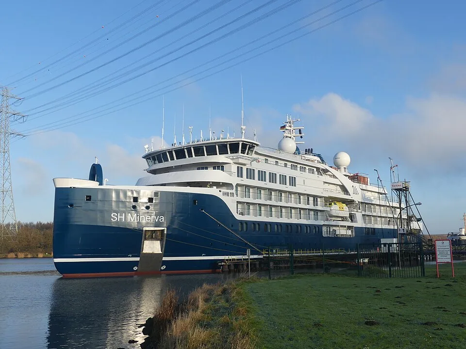 A large, blue-hulled expedition cruise ship named "SH Minerva" is docked along a calm waterway under a blue sky with scattered clouds. The ship features several levels of white upper decks, large windows, and a distinctive rounded bow. A grassy bank and power lines are visible on the shore.