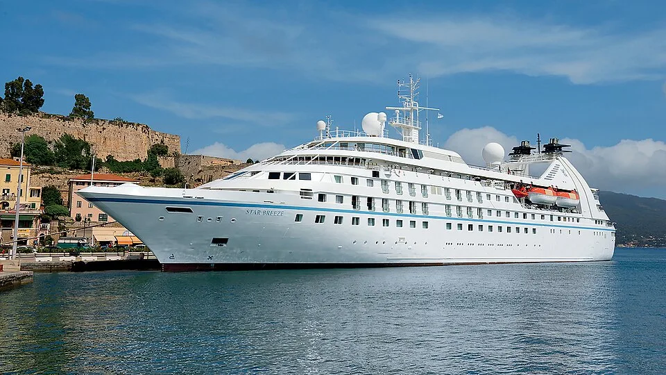 A sleek, all-white luxury cruise ship named "Star Breeze" docked in calm blue water under a bright blue sky with light clouds. A rocky, green-topped cliffside town rises in the background behind the multi-level ship.