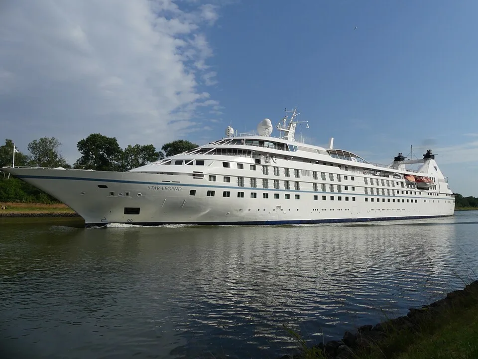 A white luxury cruise ship named "Star Legend" sailing along a calm waterway lined with green trees. The multi-deck vessel features a pointed bow, rows of windows, and dark twin smokestacks at the stern, all under a blue sky scattered with wispy clouds.