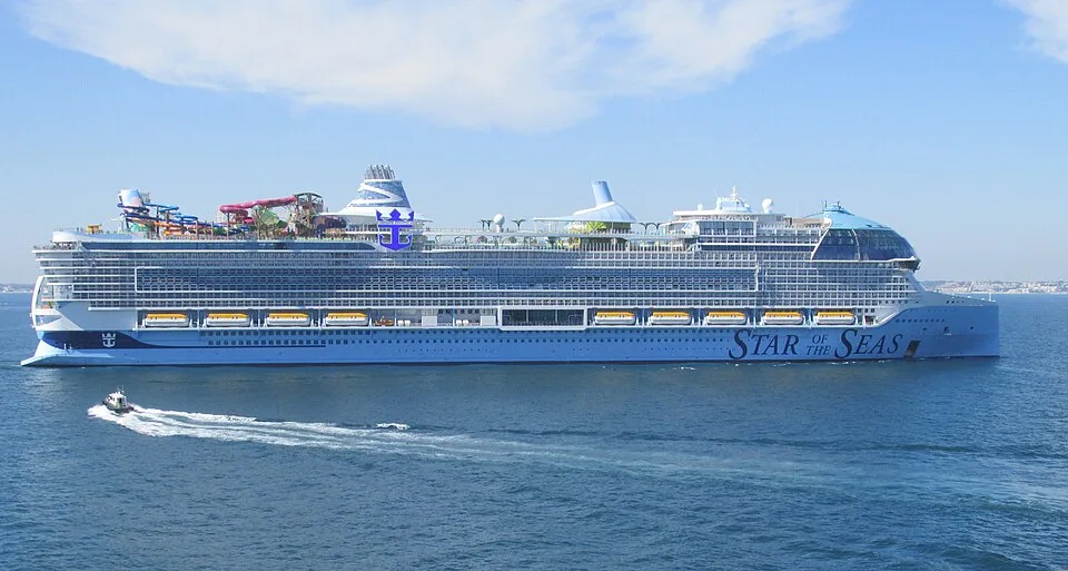 The massive Royal Caribbean ship Star of the Seas sails on blue water under a clear sky. The light-blue vessel features its name near the bow, multiple rows of windows and yellow lifeboats, colorful water slides on the top deck, and a small motorboat cruising in the foreground.