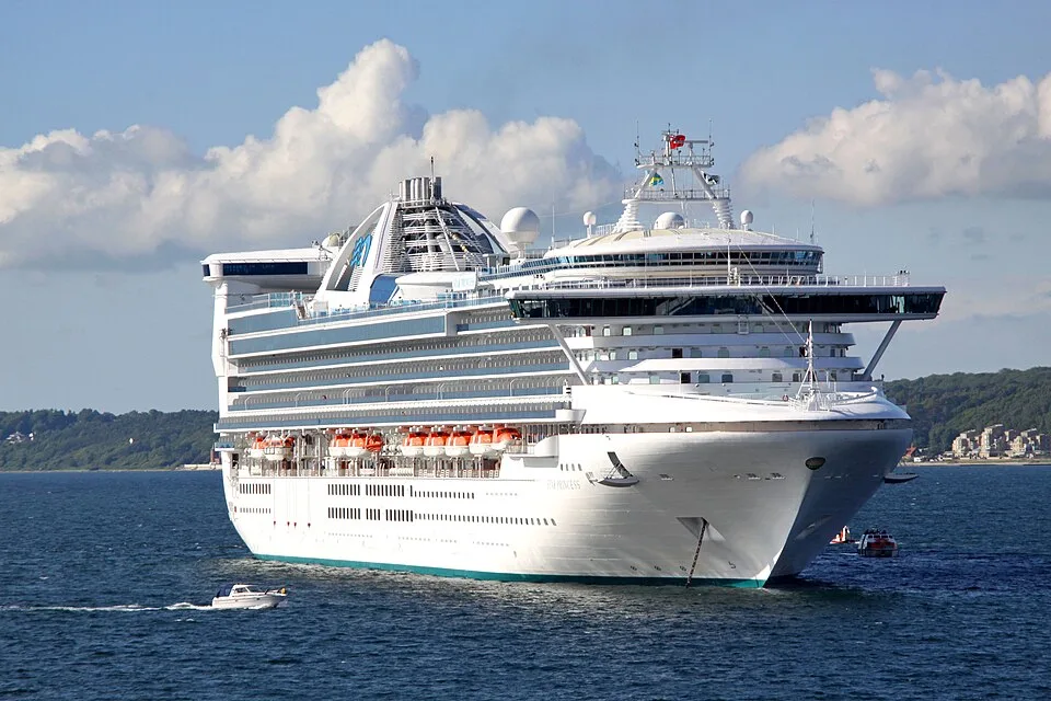 A massive white Princess Cruises ship floats on calm blue water. The vessel features a wide, multi-level glass bridge, rows of balconies, orange lifeboats along the side, and a unique blue logo on its top structure. A small white boat sails nearby.