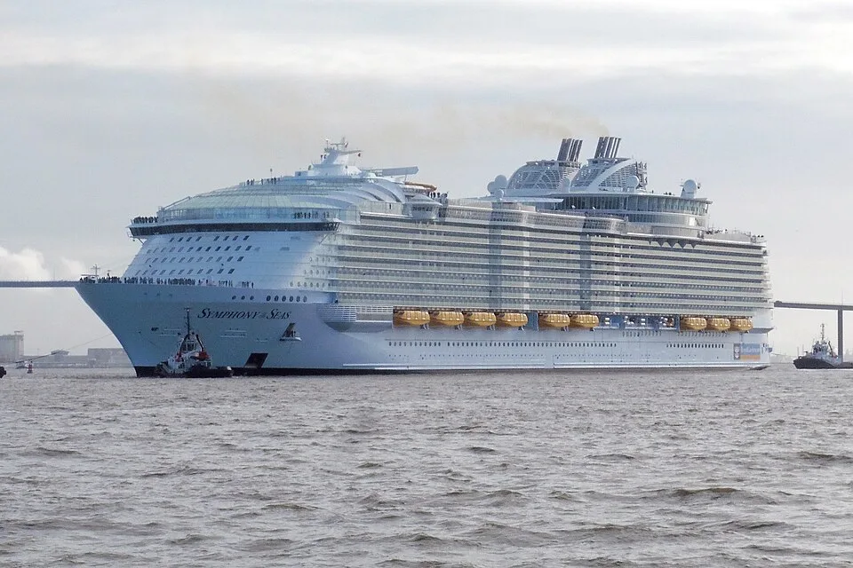 A massive white Royal Caribbean cruise ship named "Symphony of the Seas" navigating choppy water. The multi-deck vessel features a wide bow, rows of balconies, yellow lifeboats, and exhaust plumes coming from its smokestacks, with small tugboats nearby under an overcast sky.
