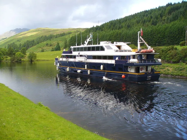 A dark blue and white passenger ship named "Lord of the Glens" navigates a narrow waterway. Its hull is lined with white fenders as it leaves a slight wake on the dark water. The scene is bordered by a green grassy bank on the left and a steep, tree-covered hill rising under a cloudy sky.