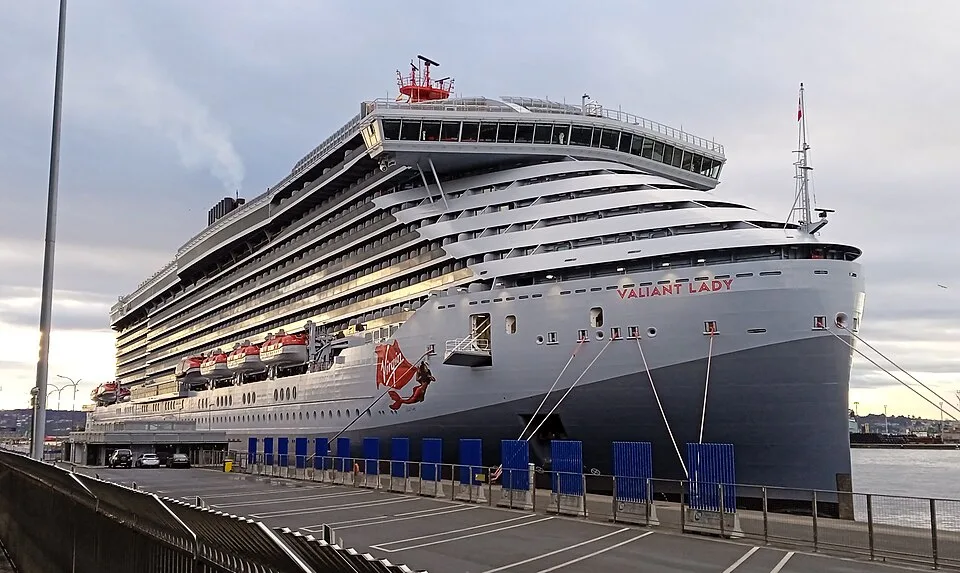 A massive grey and white Virgin Voyages cruise ship named "Valiant Lady" docked at a concrete pier. The modern vessel features a distinctive red mermaid graphic on its hull, tiered cabin decks, red accents on top, and mooring lines secured to the dock under a cloudy sky.